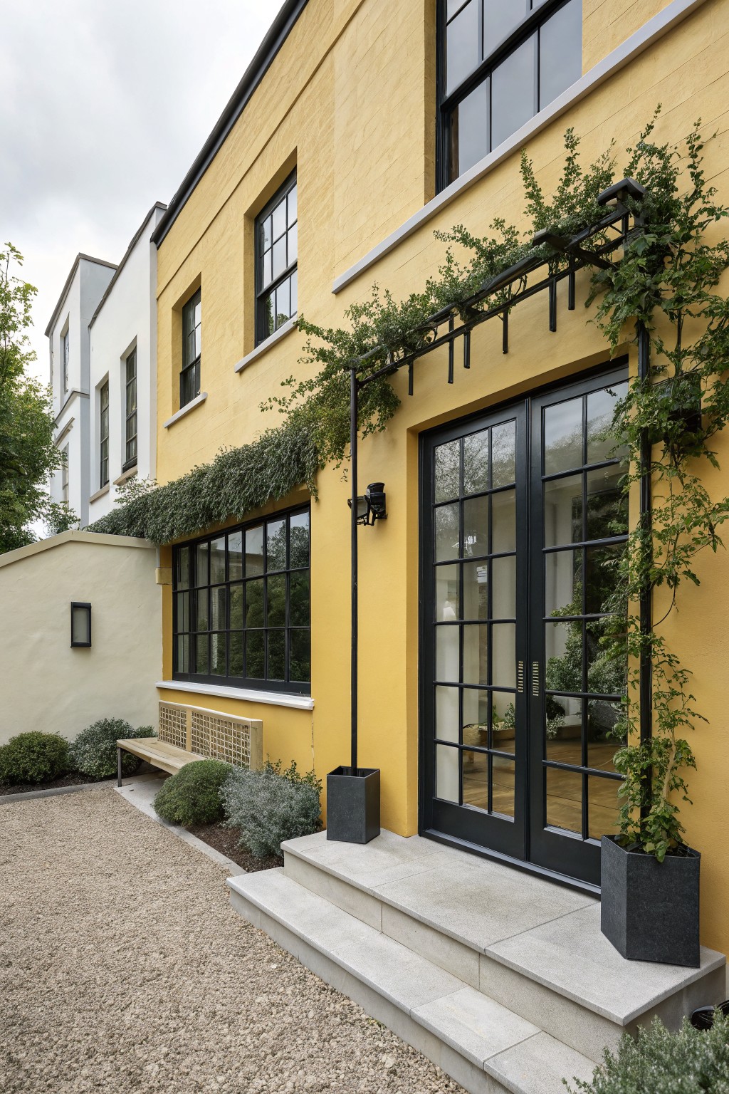 Yellow brick house exterior with black steel-framed windows and double glass doors at the entrance, climbing plants on a metal trellis overhead, concrete steps, gravel path, wooden bench, and potted shrubs in the yard.