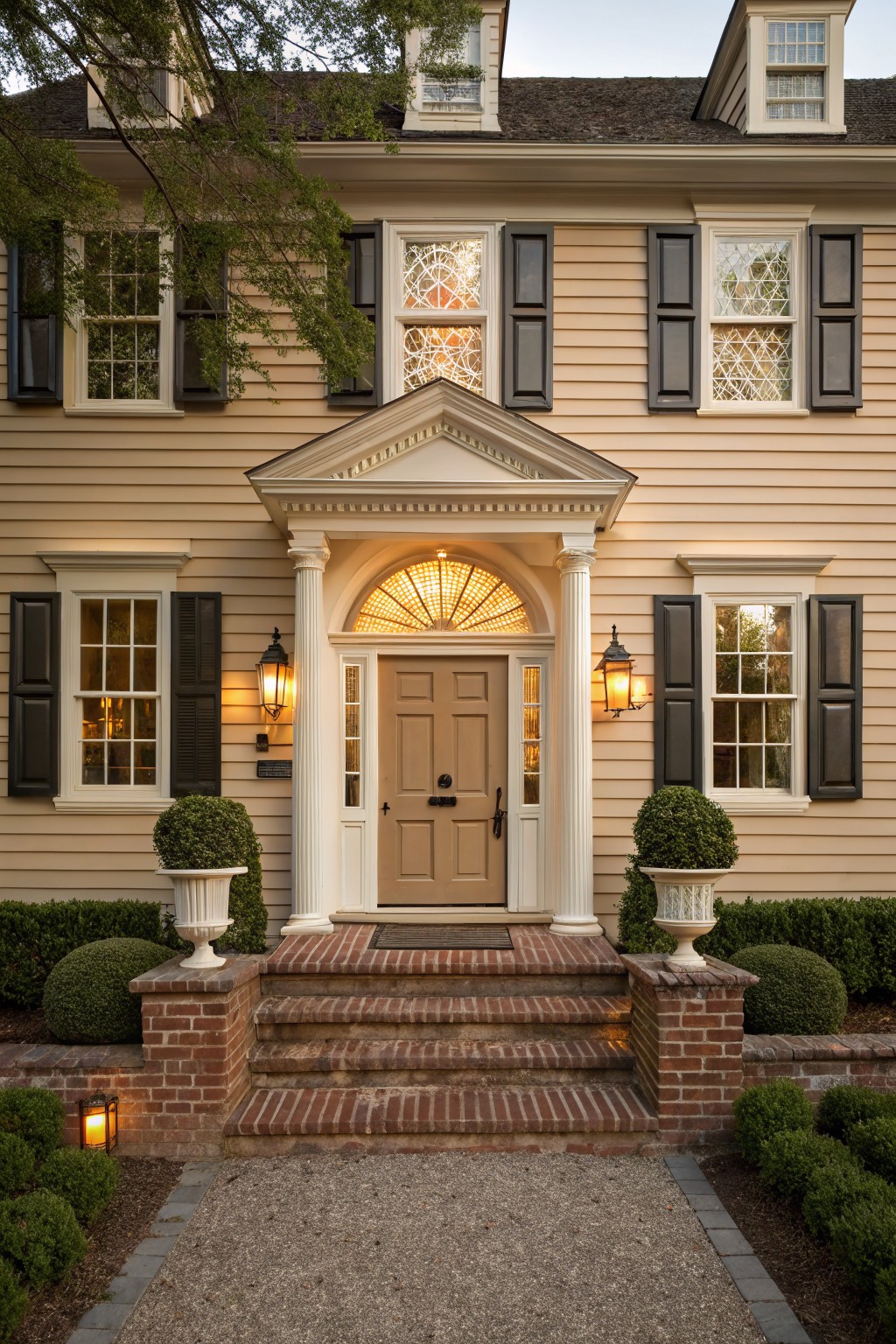 Beige clapboard two-story house with black shutters, white pedimented portico entry supported by columns, fanlight transom over a wood door, flanked by lanterns, brick steps, boxwood topiaries, and gravel path with surrounding shrubs.