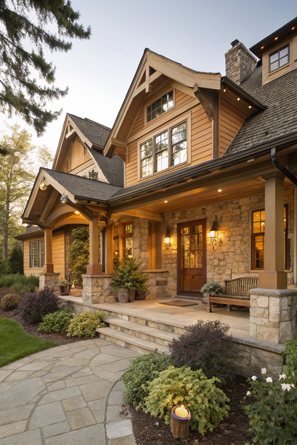 Two-story house exterior with light brown horizontal wood siding, dark shingled roof, covered front porch with exposed timber beams and stone pillars supporting it, double wood front doors, flanked by large windows, stone base, steps leading to porch, surrounded by shrubs, plants, and stone pathway.
