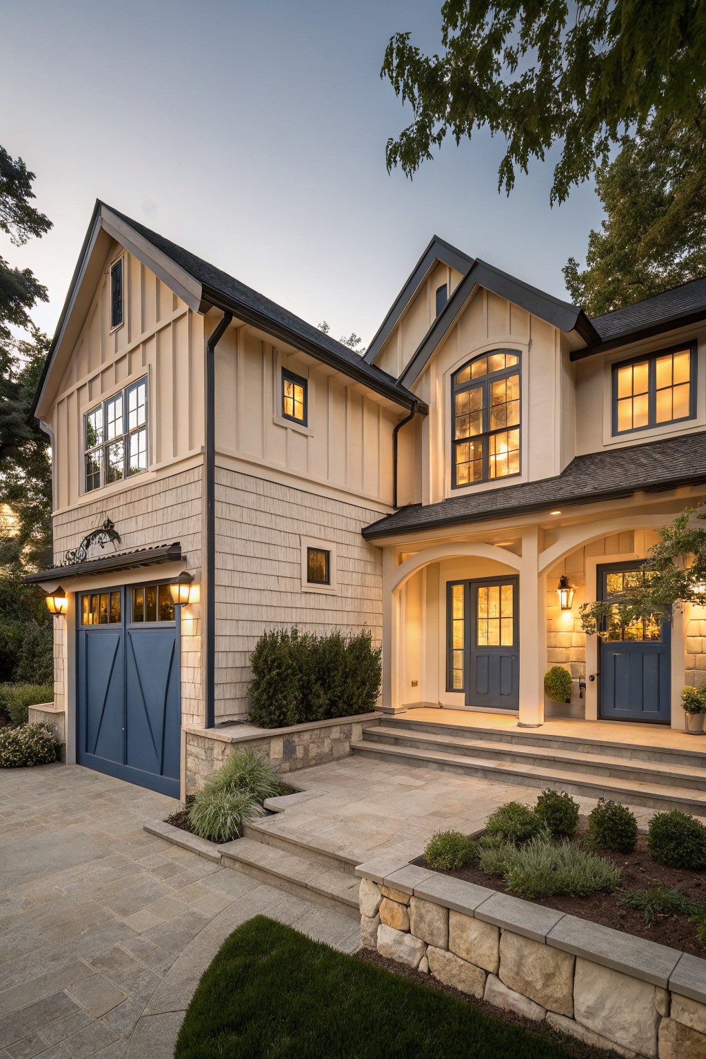 Two-story house exterior with light beige board-and-batten and shingle siding, navy blue garage doors and entry doors under an arched portico, stone retaining wall, steps, and low plantings at dusk.