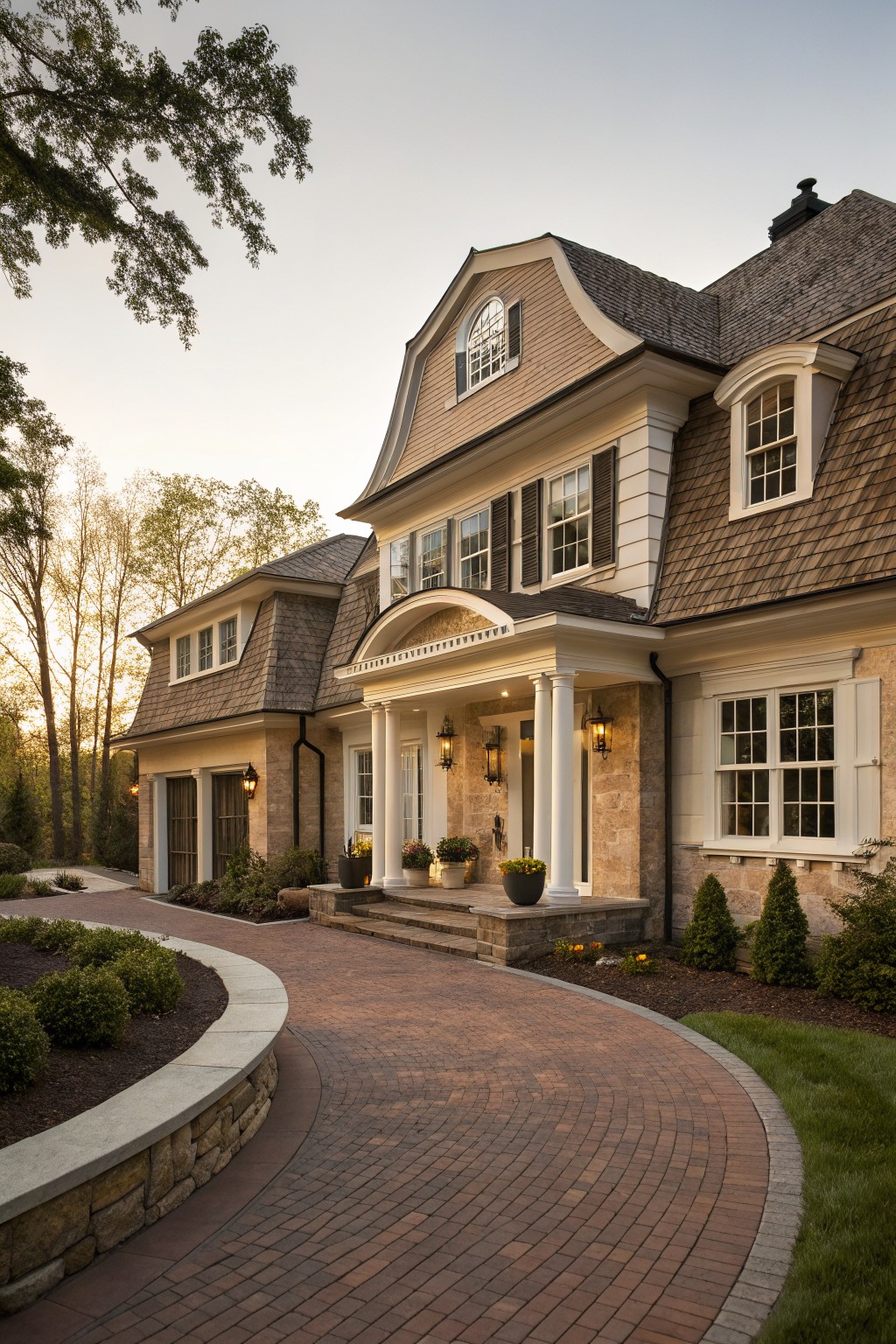 A two-story house with light brown shingle siding and roof, beige stone base, white columned front porch, brick curved driveway, low stone walls, shrubs, and trees in soft evening light.