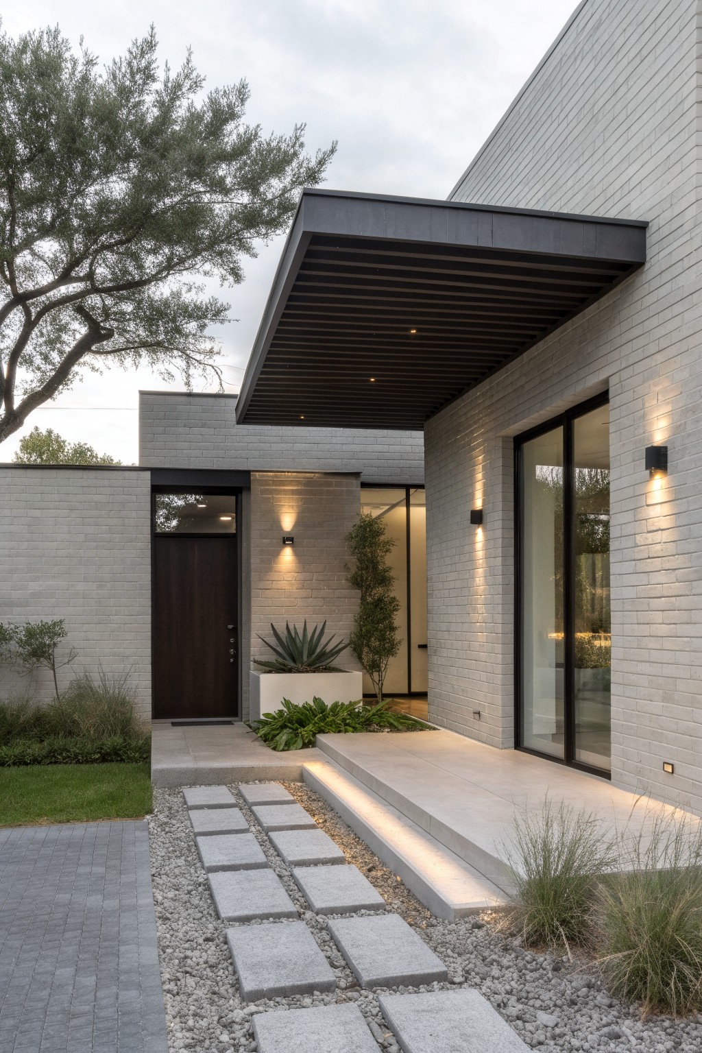 Contemporary gray brick house exterior with dark wood front door under black cantilevered overhang, large glass window beside it, stone step path with integrated lighting, and sparse desert landscaping including agave and grasses.