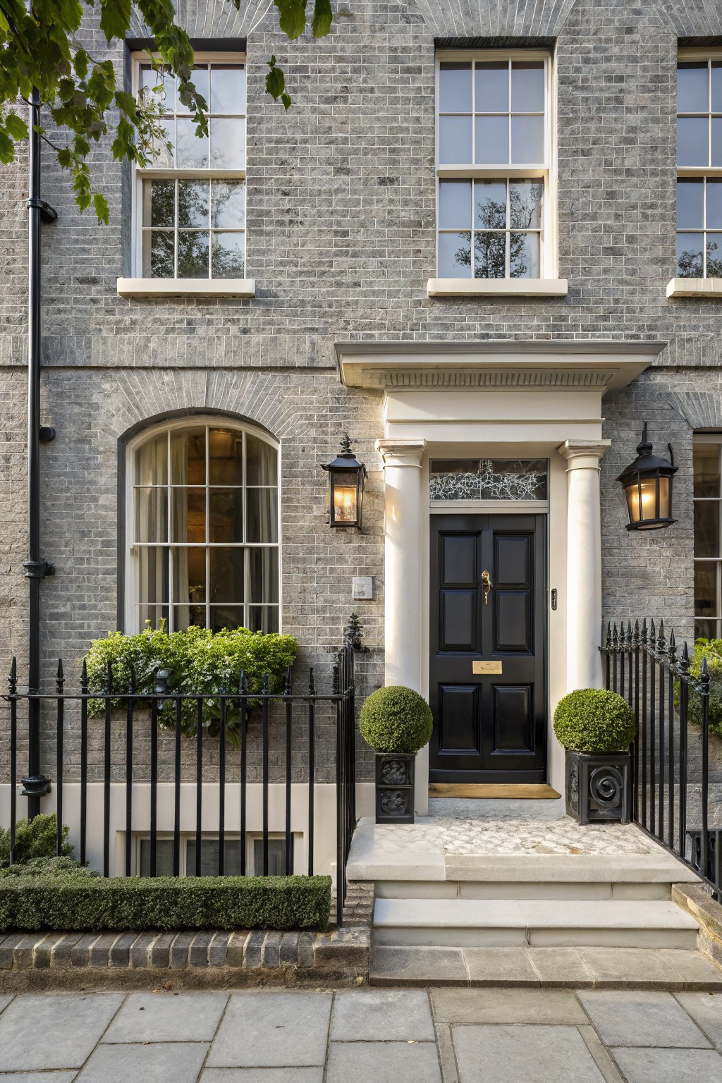 Gray brick townhouse exterior with arched windows, black double front door under a white portico supported by columns, iron railings with topiary plants, and lanterns on either side.