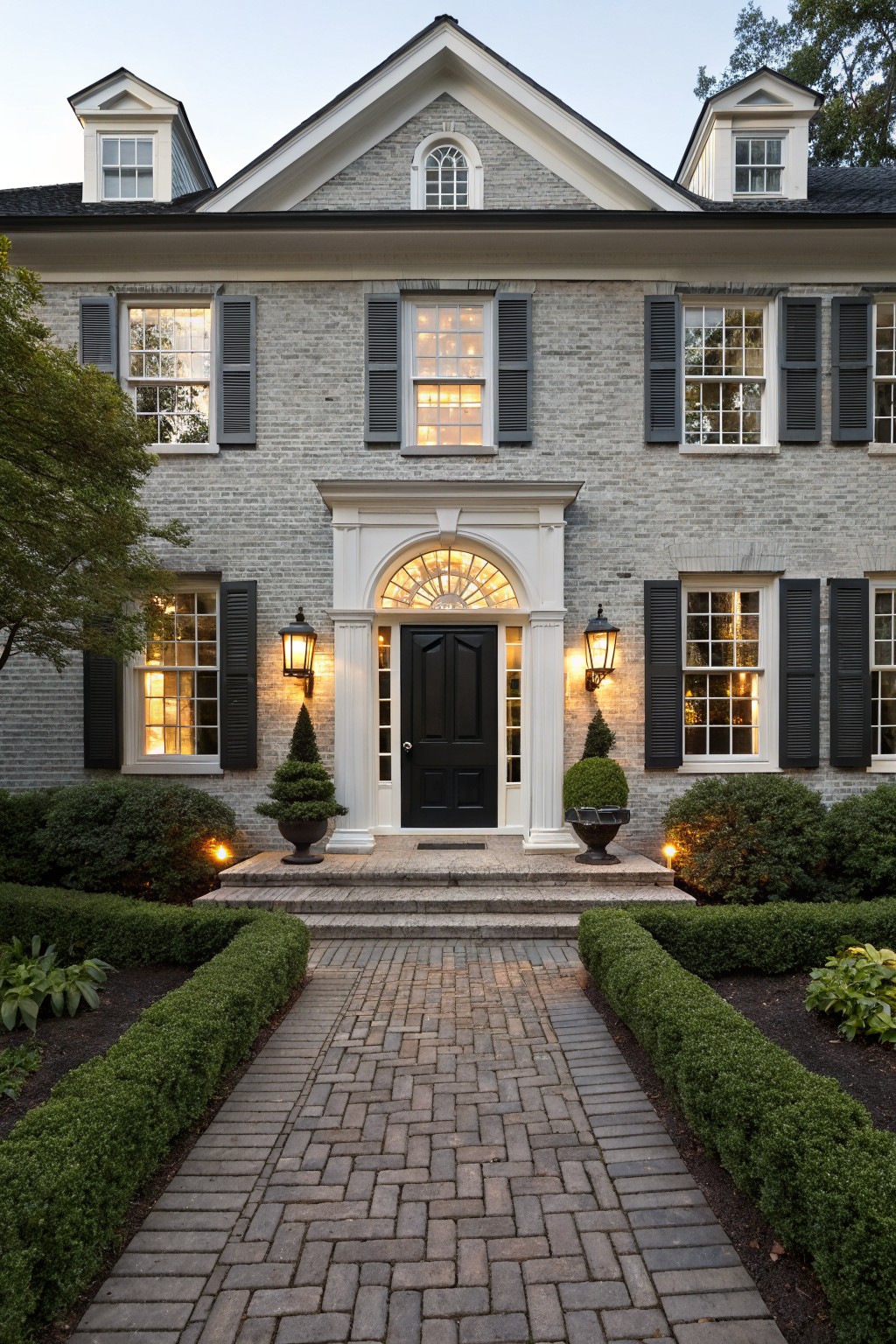 Front view of a two-story gray brick house with white trim, black shutters on multi-pane windows, arched entry with black door flanked by lanterns and topiaries, boxwood hedges, and a brick walkway.