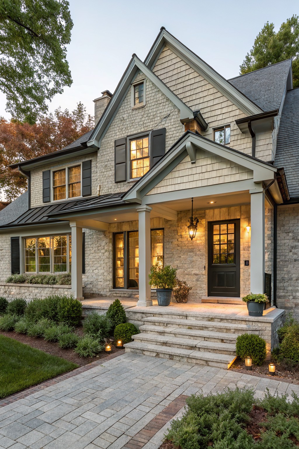 Front exterior of a two-story house with textured gray brick walls, wood shingle gables, covered porch supported by columns, black front door, large multipane windows with black shutters, stone steps, potted plants, and brick pathway bordered by shrubs and lawn.