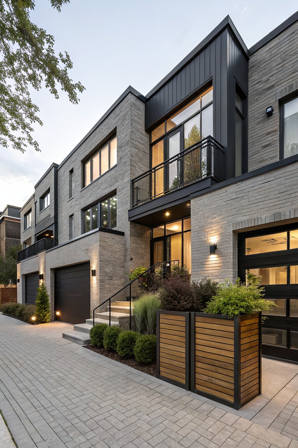 Multi-level townhouse exterior with textured gray brick walls, black metal cladding on upper sections and balcony railing, large windows, double black garage doors, front entry stairs with plants, wooden planters, and paver walkway at dusk.