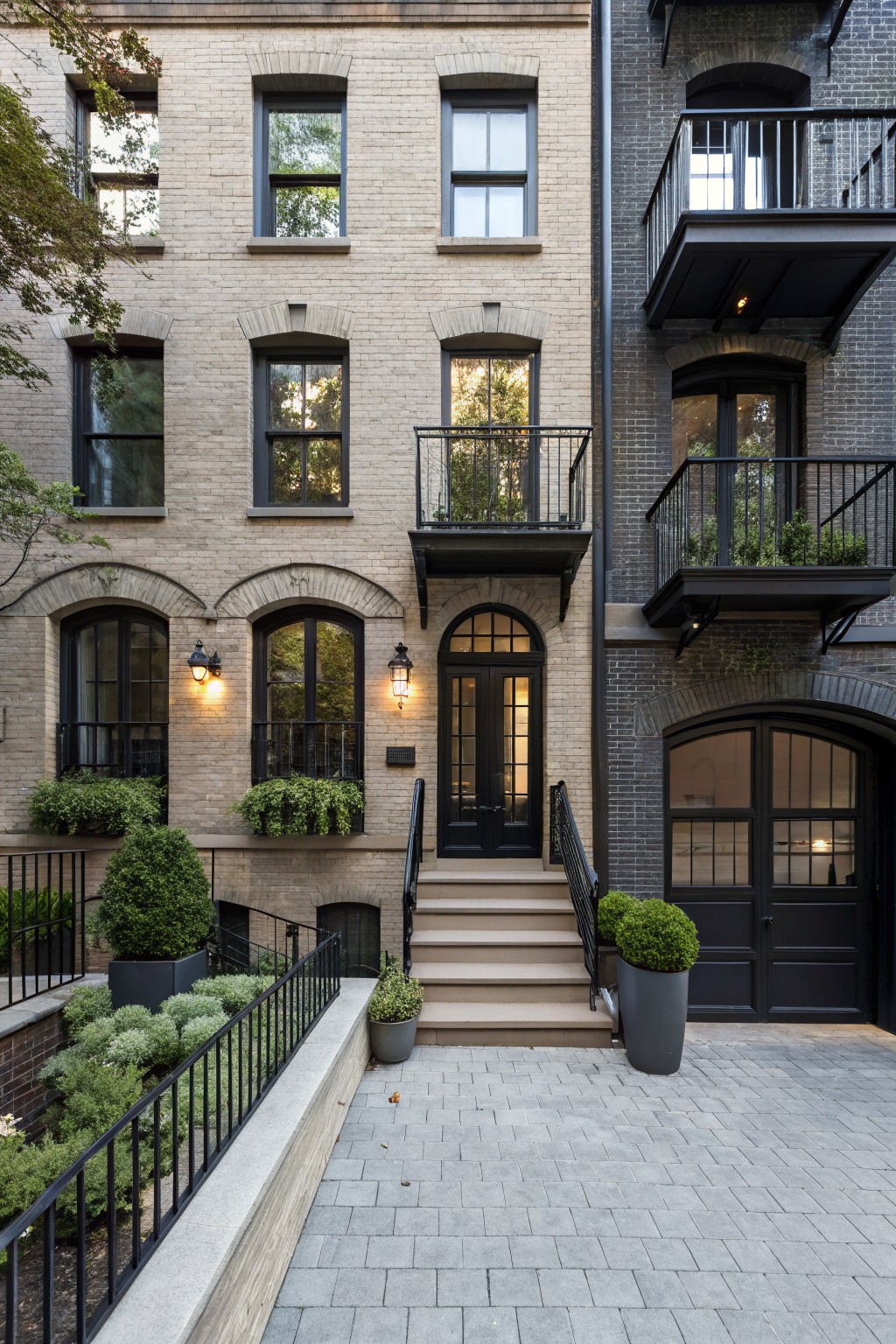 Two adjacent townhouse facades, one in light beige brick with arched windows, black lanterns, and a front door entry with steps, the other in dark gray brick with a metal balcony, arched garage door, and potted plants, set against a paved courtyard with low shrubs and black metal railings.