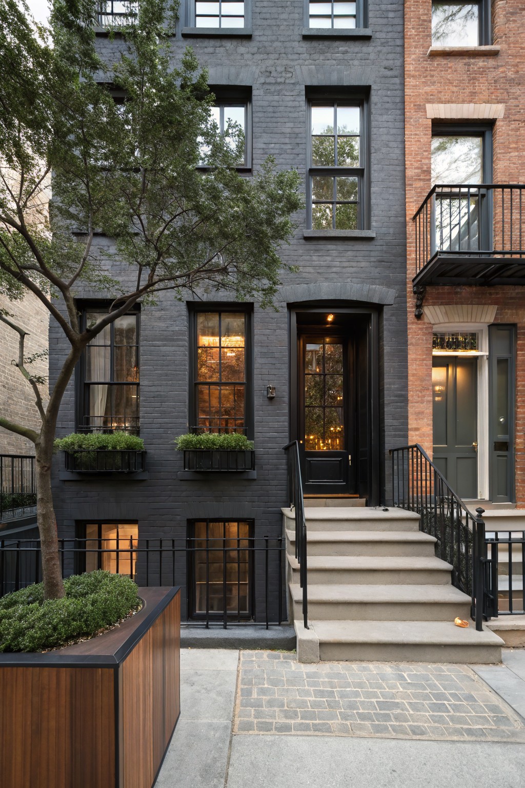 Dark gray brick two-story townhouse with black-framed multipane windows, window boxes of greenery, black front door, concrete entry steps with black railing, large wooden planter box with tree and shrubs beside it, adjacent to red brick house on cobblestone street.
