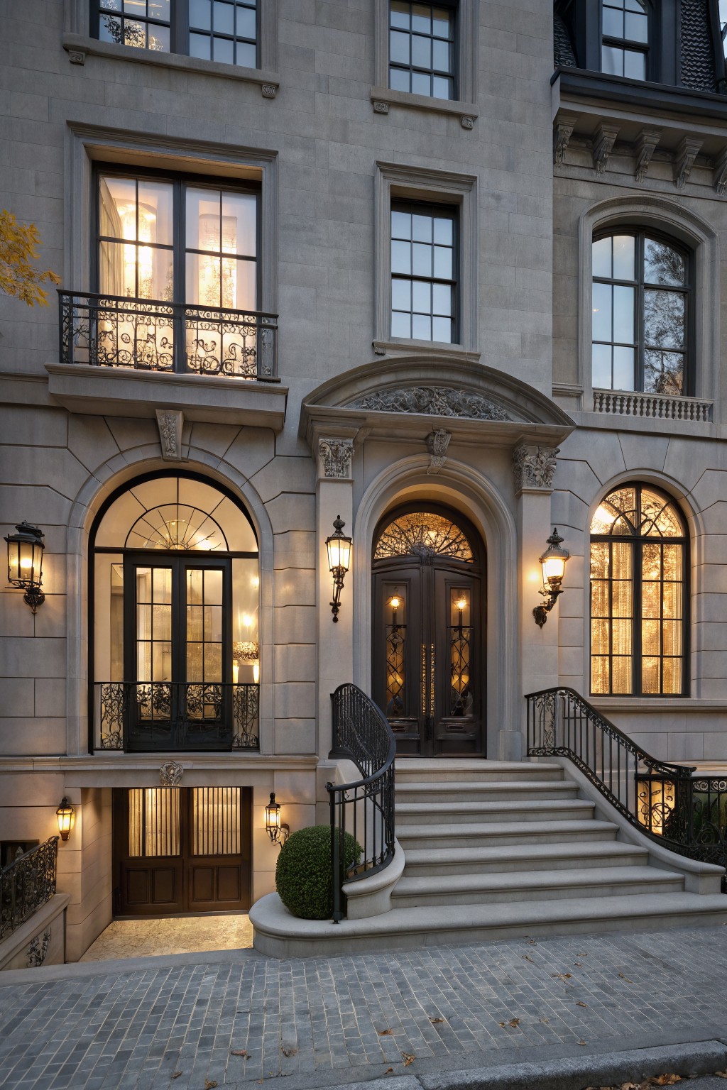 Multi-story gray limestone townhouse exterior at dusk with black-framed arched windows and doors, curved stone entry stairs with wrought iron railings, lanterns, and a lower garage door.