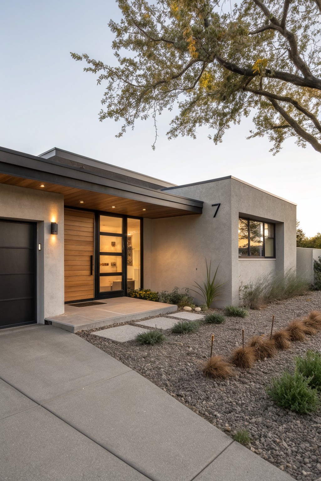 Front exterior of a modern single-story house with light gray stucco walls, black garage door, vertical wood slat front door with glass panels, black window frames, and gravel landscaping with grasses and rocks.