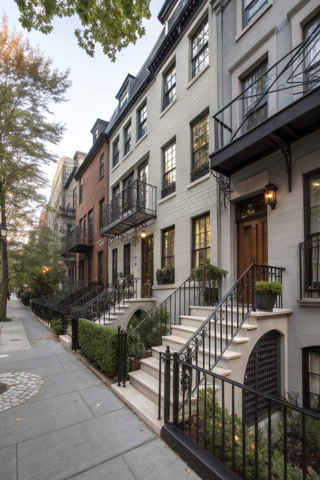 Row of attached gray brick and stucco townhouses with black wrought iron balconies, stair railings, and entrance gates on a sidewalk lined with trees and shrubs.