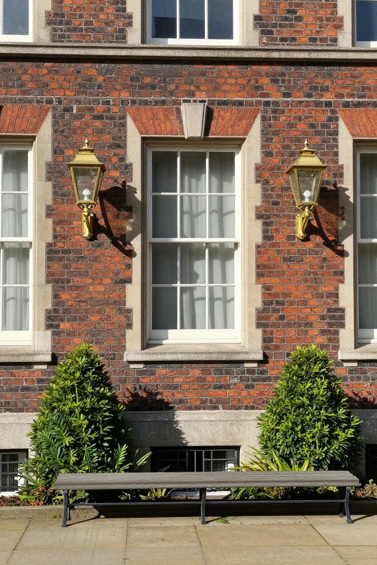 Red brick house exterior with three sets of multi-pane sash windows including projecting bays, flanked by ornate gold wall lanterns, boxwood topiaries on either side, and a wooden bench on a paved courtyard.