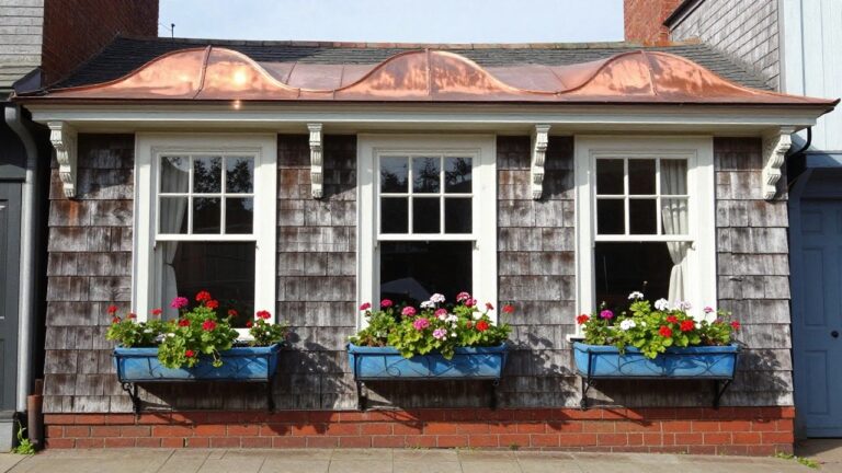Close-up exterior view of an octagonal bay window on a shingled house corner, featuring white-framed triple windows, copper roof accents, a blue cushioned bench seat below, and black metal planters with red geraniums on brick supports.