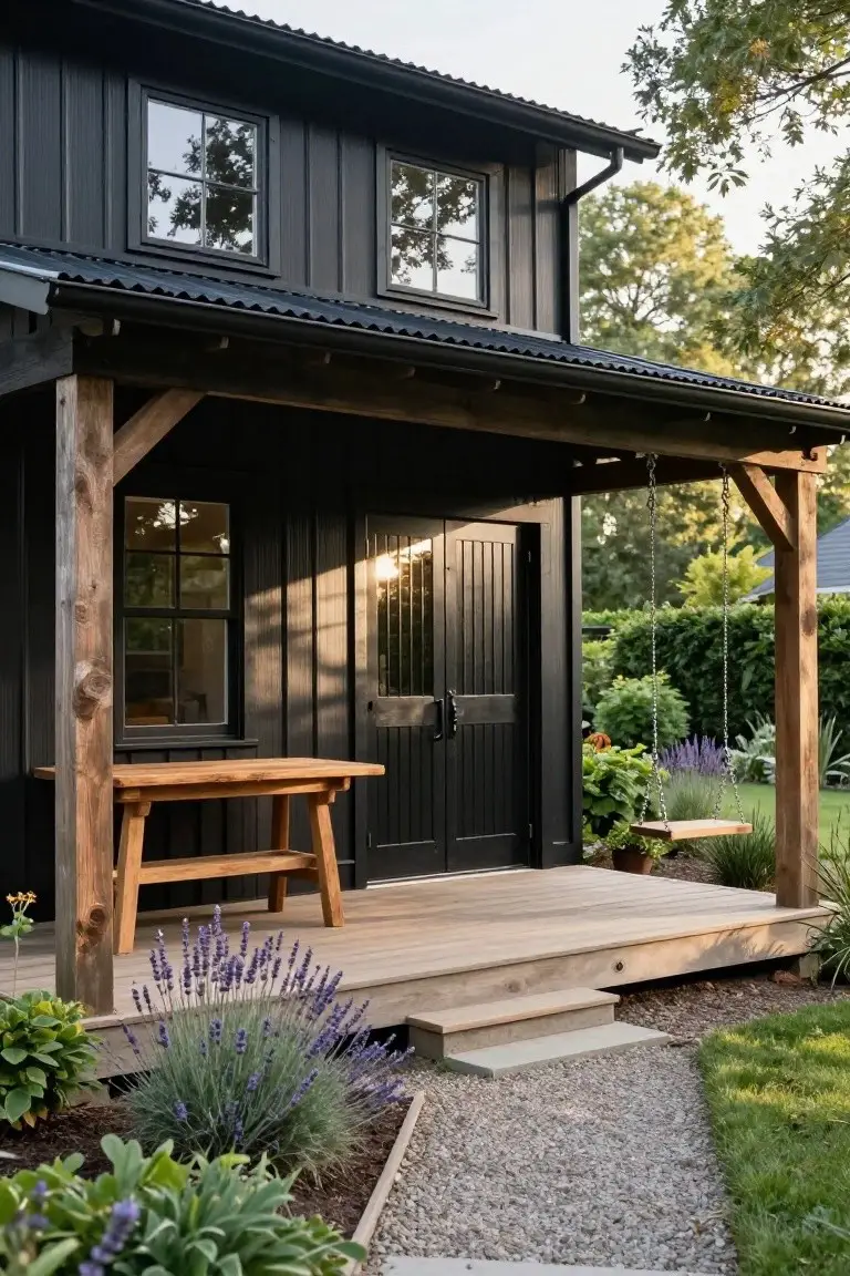 Black board-and-batten barndominium exterior with covered porch on timber posts, wooden table and swing on deck, double black doors, lavender plants, gravel path, and grass lawn.
