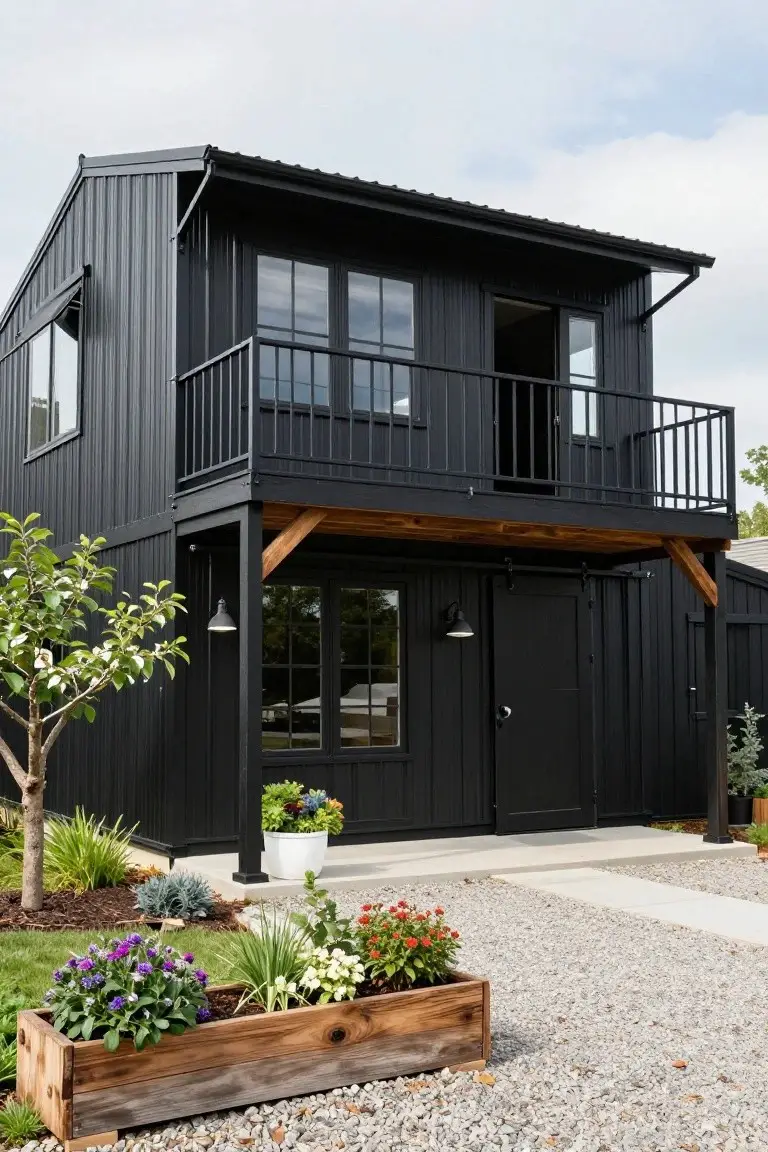 Two-story black barndominium with corrugated metal siding, wooden balcony and beams, large windows, sliding barn door, and gravel driveway with plants and wooden planter box.