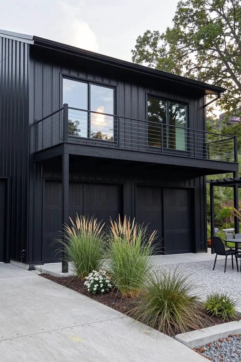 Black barndominium exterior featuring corrugated metal siding, double garage doors, overhanging balcony with cable railings, large windows, and gravel landscaping with ornamental grasses.