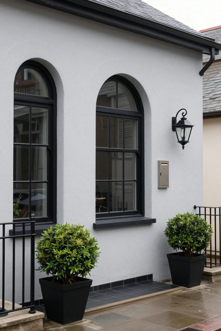 White stucco house exterior with two black-framed arched windows, black wall lantern, metal mailbox, potted boxwood shrubs in black containers, and black metal railing next to a paved entry path.