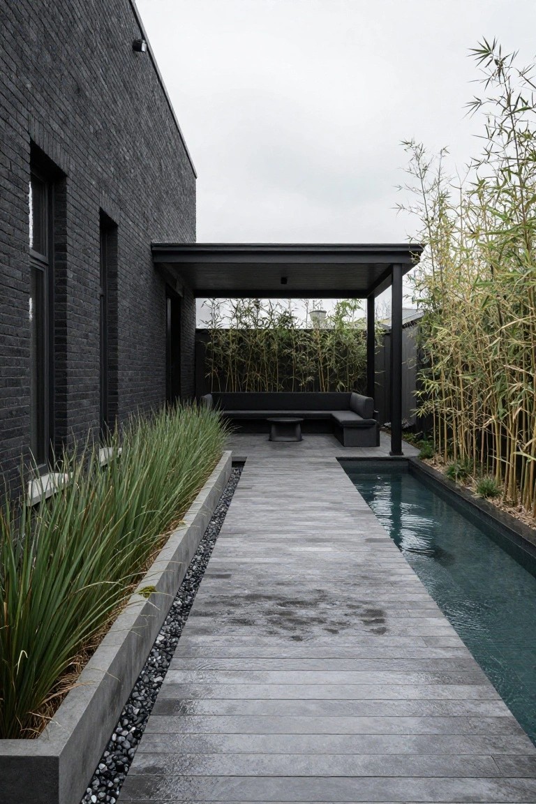 Side view of black brick house wall with a gray wooden deck pathway running parallel to a narrow rectangular pool, bordered by grasses, pebbles, and bamboo screens leading to a covered black seating area.