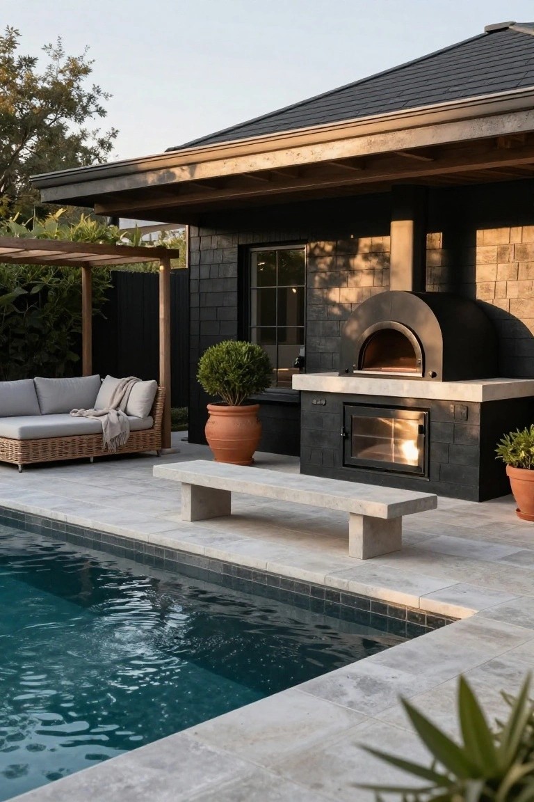 Black dome-shaped wood-fired pizza oven and fireplace built into a dark brick masonry unit on a tiled poolside patio next to a dark shingled house exterior, with a concrete bench, lounge seating, potted plants, and pool in the foreground.