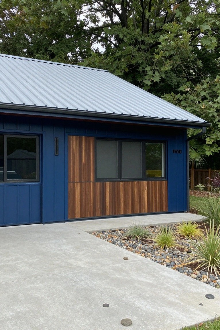 Side exterior of a house with navy blue vertical siding, a section of light brown vertical wood cladding beside tinted windows, gray metal roof, concrete pad, and gravel garden bed with grasses.