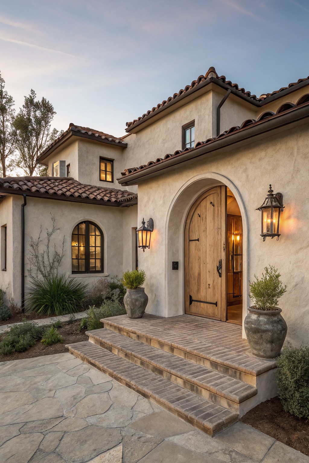 Gray stucco house exterior with terracotta tile roof, tall arched wooden front door, wrought iron lanterns, large terracotta pots with plants, brick entry steps, and flagstone walkway at dusk.