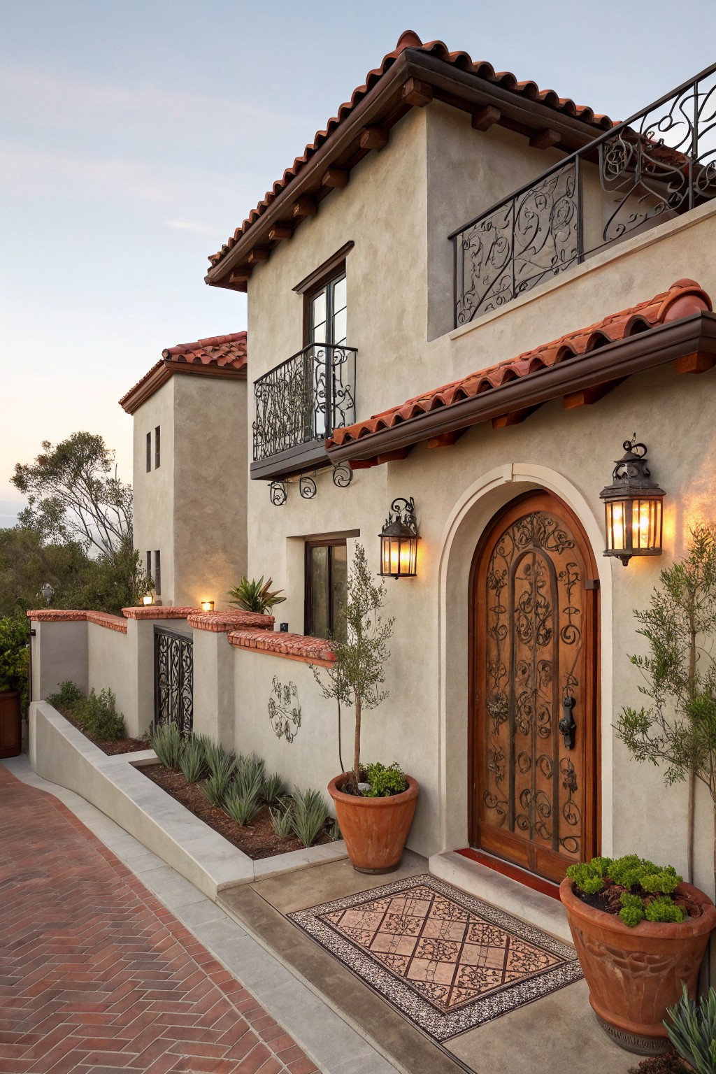 Gray stucco house exterior with red tile roof, wrought iron balcony and gate, arched wooden entry door with metal knocker, paired wall lanterns, potted plants, and brick pathway.