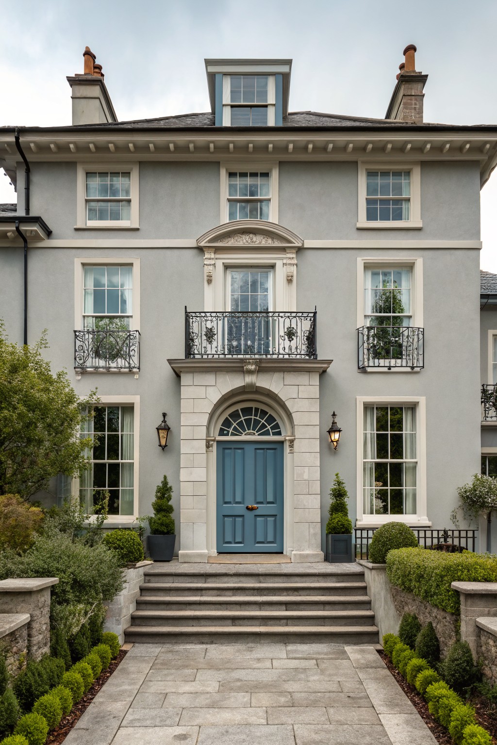 Two-story gray stucco house exterior with symmetrical sash windows, wrought-iron balconies, turquoise blue arched front door in stone surround, wall lanterns, stone steps, and formal evergreen landscaping.