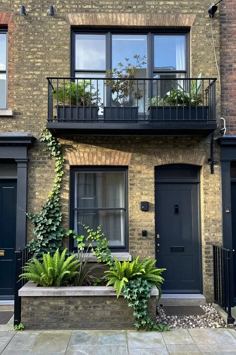Yellow brick two-story terraced house exterior with black-framed windows and double door, ivy climbing the left side from neighboring black door, ferns and plants in a stone trough planter by the entry steps, black metal balcony above with potted plants, and gravel mulch on the doorstep.
