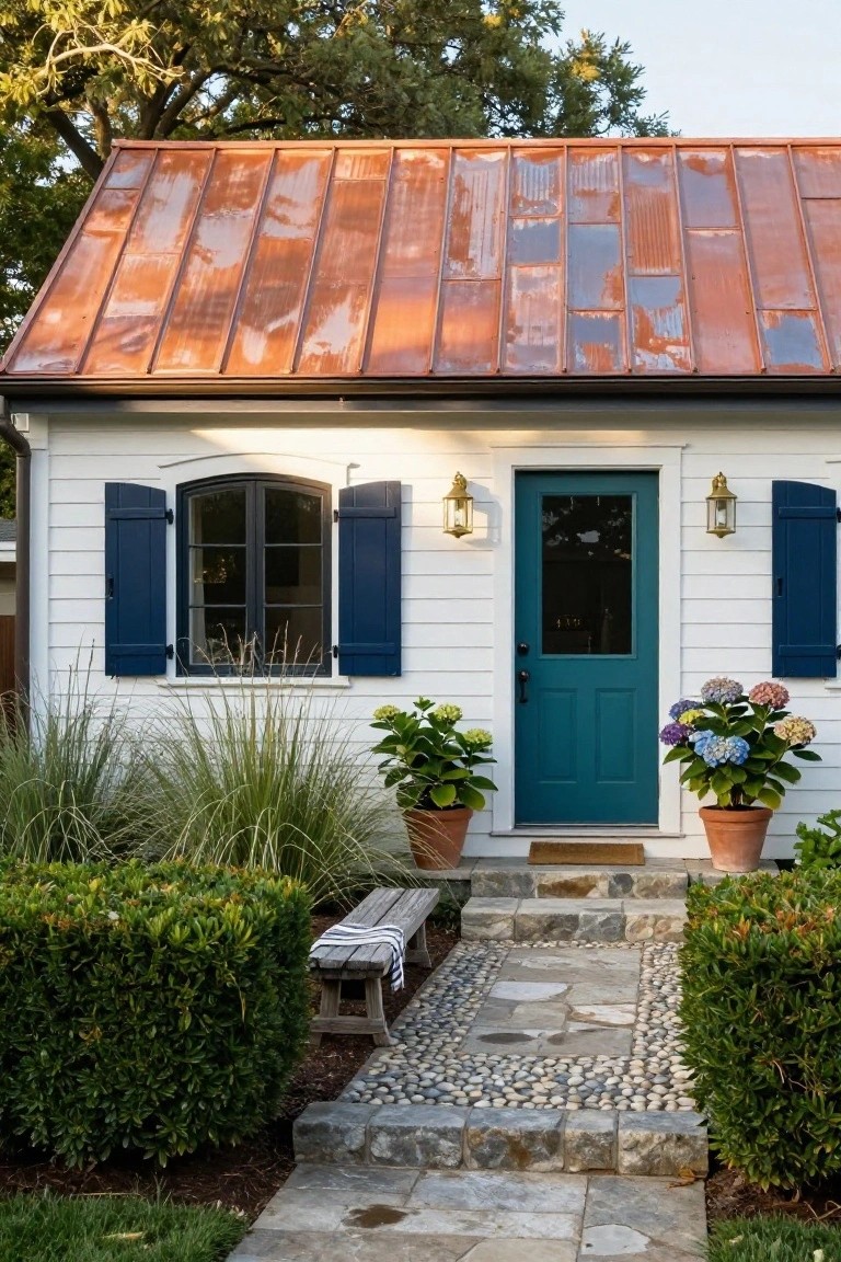 Small white clapboard house with patinated copper standing-seam roof, navy blue shutters on side windows, teal front door, potted hydrangeas, wooden bench, stone steps, and pebble path in front yard.