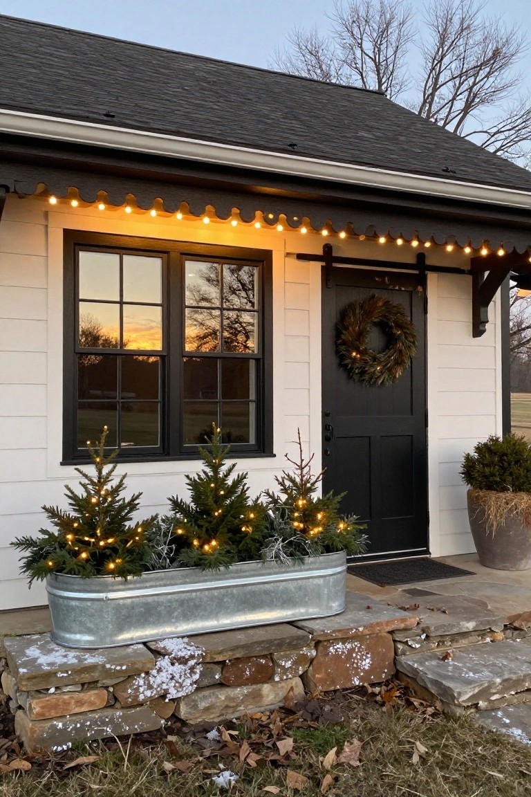 White clapboard cottage exterior with black sliding barn door, lighted mini Christmas trees in a galvanized trough planter beside the entry, string lights along scalloped eaves, evergreen wreath on door, and stone steps at sunset.