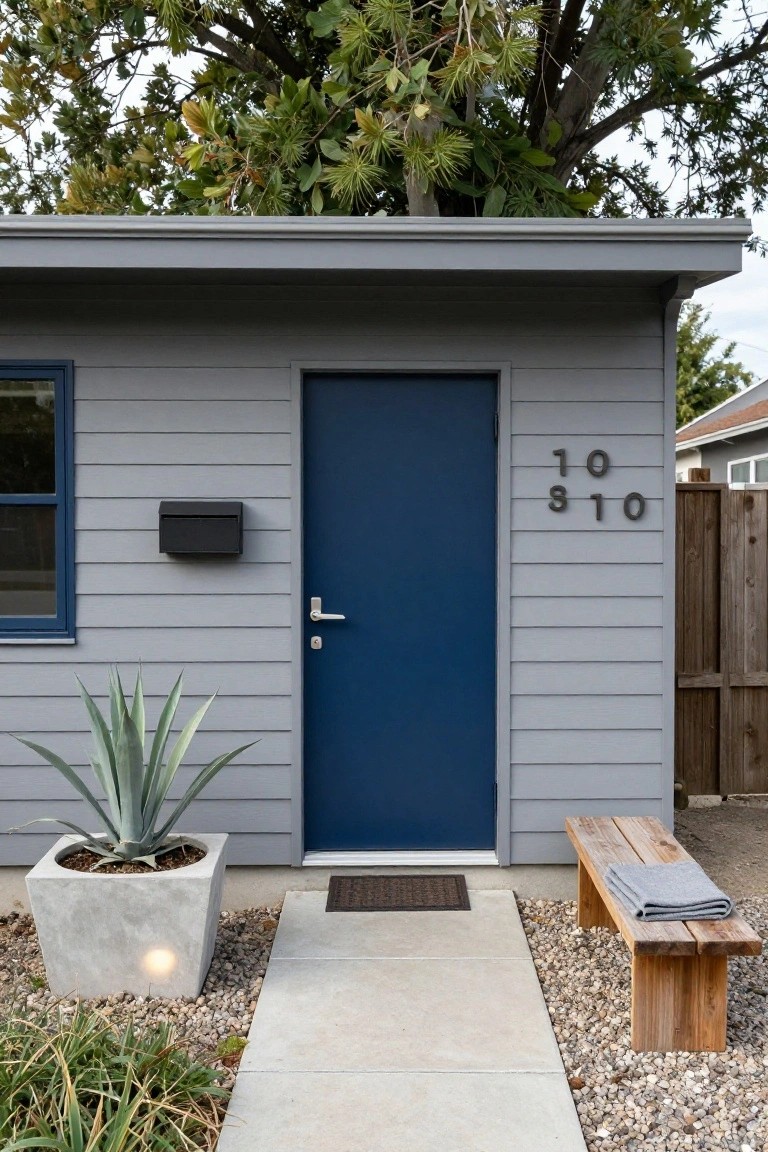 Gray clapboard house with navy blue front door numbered 810, blue window, black mailbox, potted agave plant in concrete container, wooden bench with gray blanket, concrete walkway, and gravel ground cover.