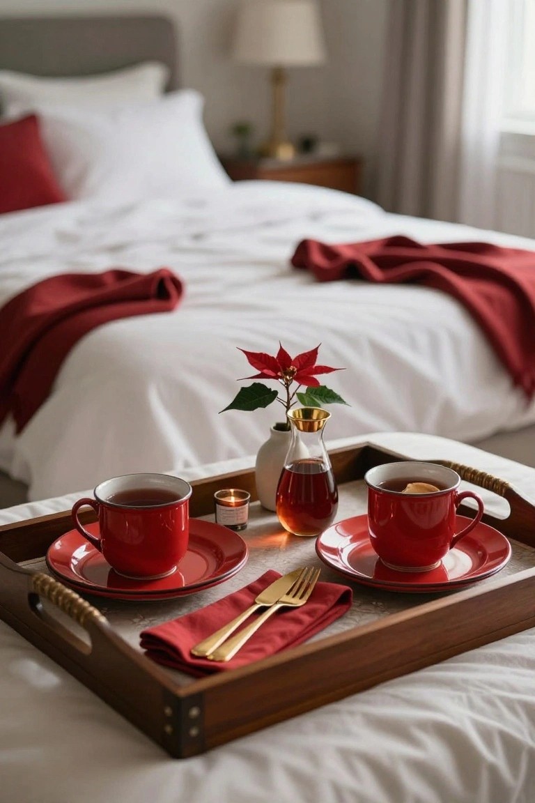 Wooden tray on white bed sheets holding two red enamel teacups with tea and orange slices, red saucers, gold knife and fork on red napkins, small lit candle, golden syrup dispenser, and potted red poinsettia plant in cozy bedroom.
