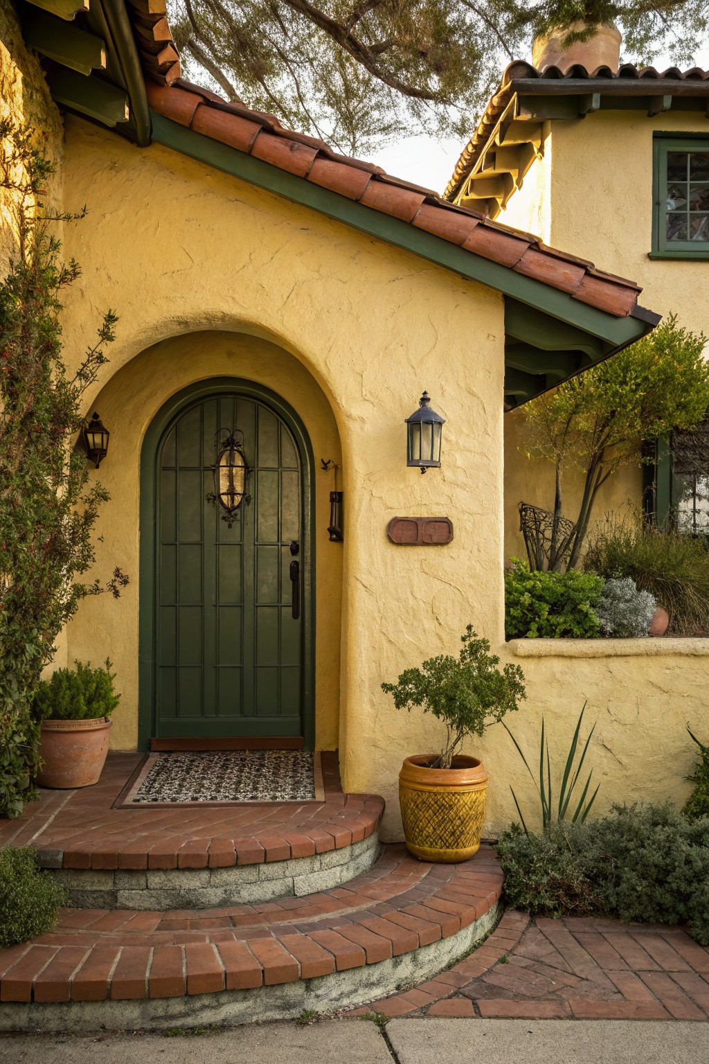 Soft yellow stucco house exterior with dark green arched wooden front door, flanked by black lanterns, red brick steps, entry mat, and surrounding potted plants and shrubs.