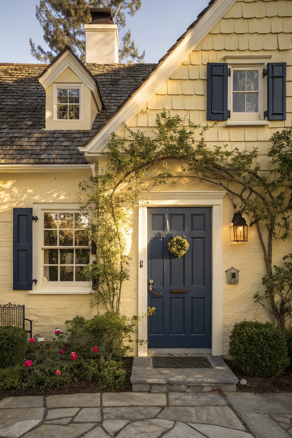 Pale yellow shingle-style house exterior featuring a navy blue front door with wreath, flanked by climbing vines and windows with navy shutters, stone steps and pathway at the entrance.