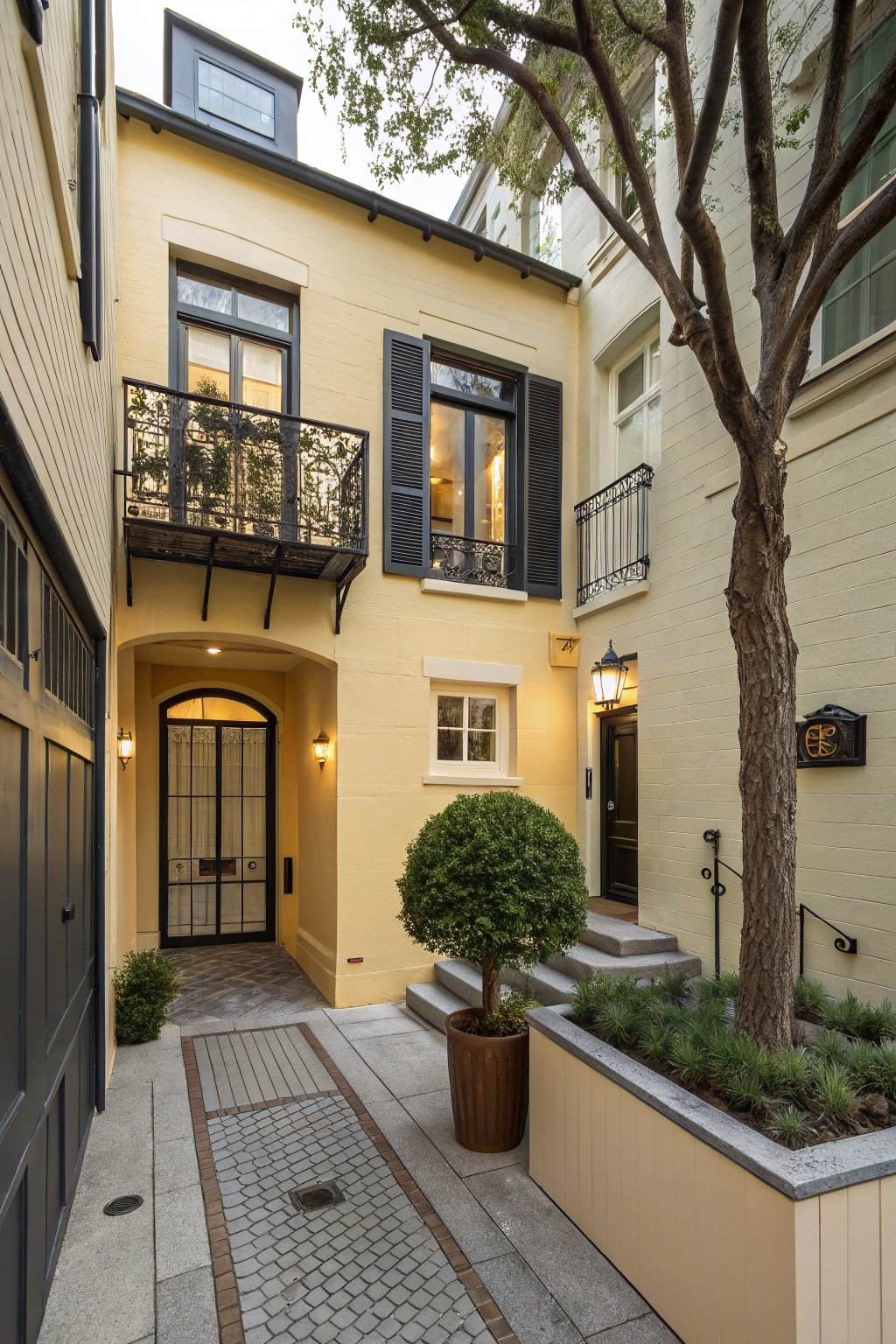 A narrow two-story pale yellow house exterior with black garage door, arched entryway featuring a glass door and lanterns, wrought iron balcony, shutters on windows, stone pathway, potted plants, and a large tree in an urban courtyard setting.