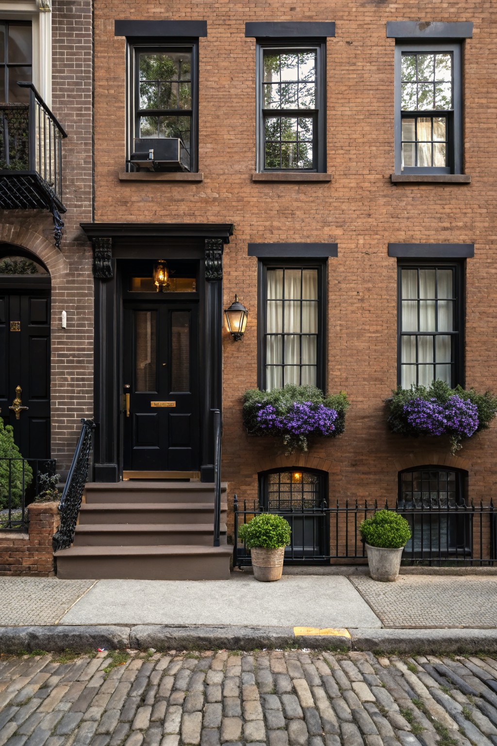 Three-story brown brick townhouse with black window frames, black front door, purple flower boxes on windows, potted shrubs, and iron railings on a cobblestone street.