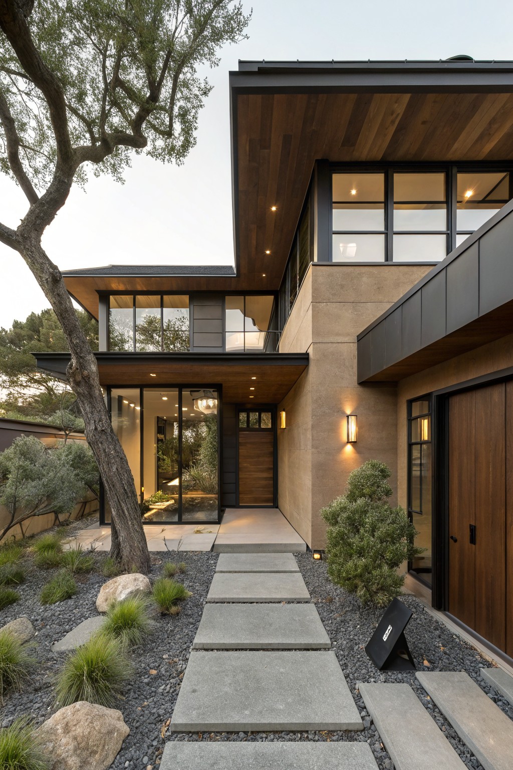 Modern two-story house exterior with brown stucco walls, black metal window frames and roof edges, cantilevered wooden overhang, wooden entry door, and stone pathway through gravel landscaping with trees and shrubs.