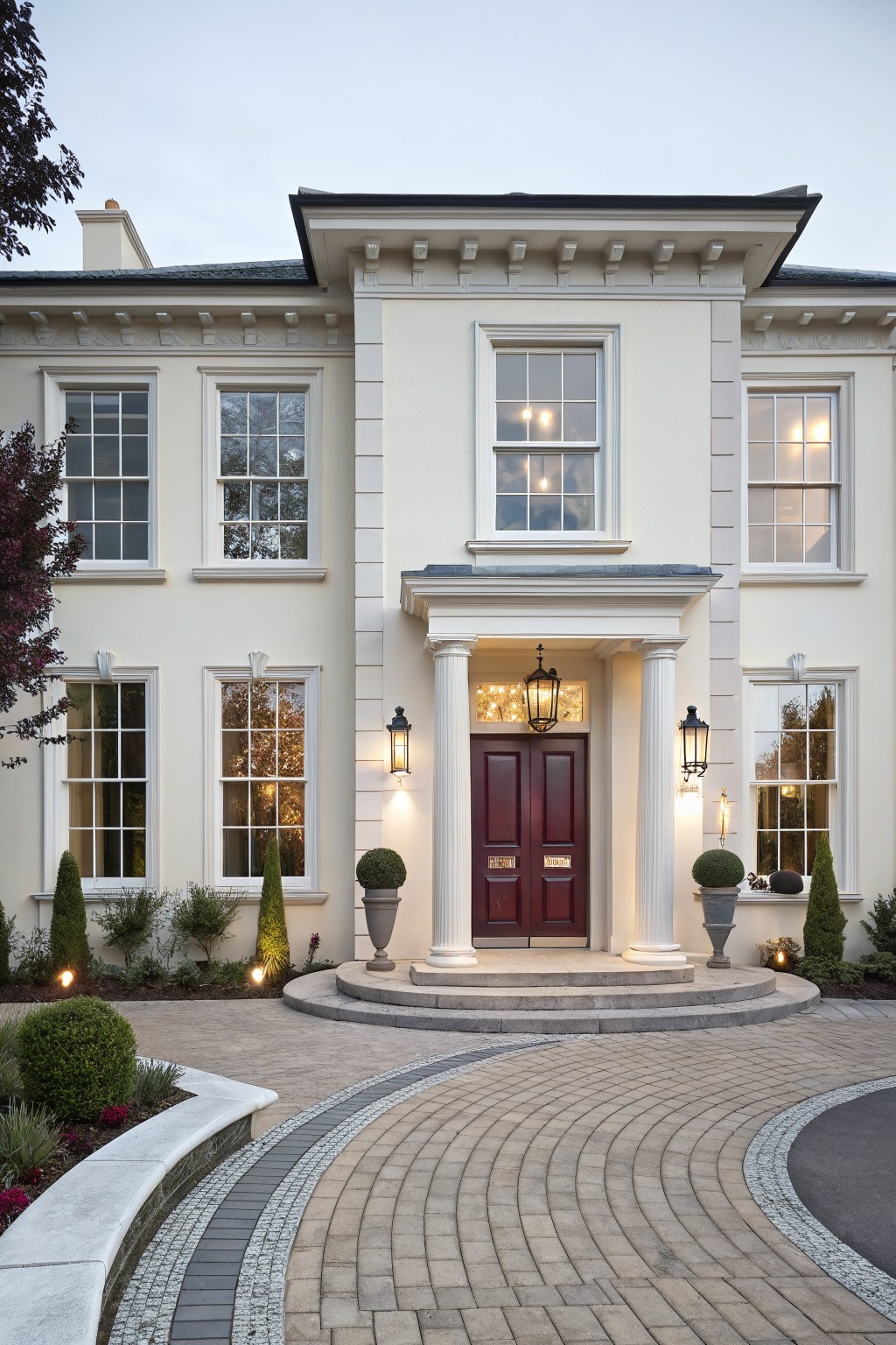 Two-story white house exterior with burgundy double front doors under a portico supported by columns, flanked by lanterns, tall windows, and landscaped driveway with curved paving.
