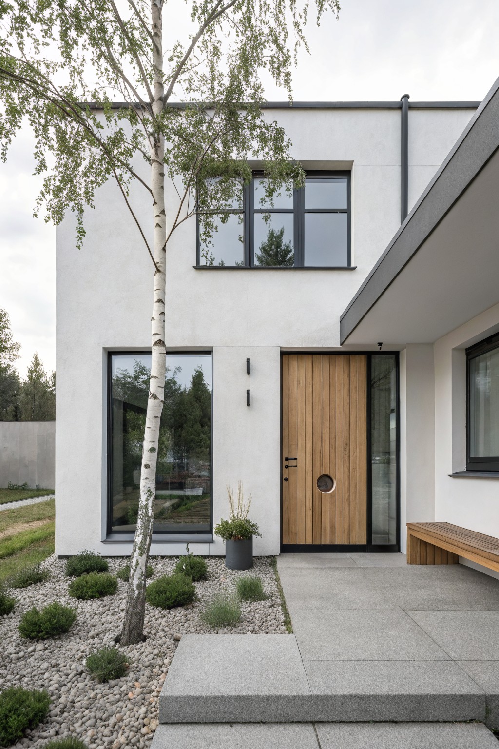 Modern white stucco house exterior featuring a tall birch tree next to a vertical wood plank front door with round window, flanked by slim wall lights, concrete entry path, gravel garden bed with low plants, and wooden bench.