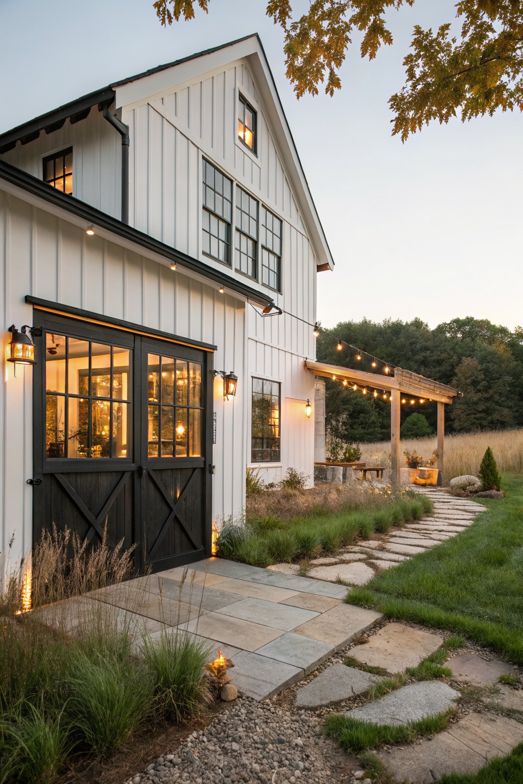 White board-and-batten barn-style house exterior featuring large black sliding garage doors with glass panels, black-trimmed windows, flanked by lanterns and landscaping with ornamental grasses, a stone pathway, and a pergola with string lights at dusk.