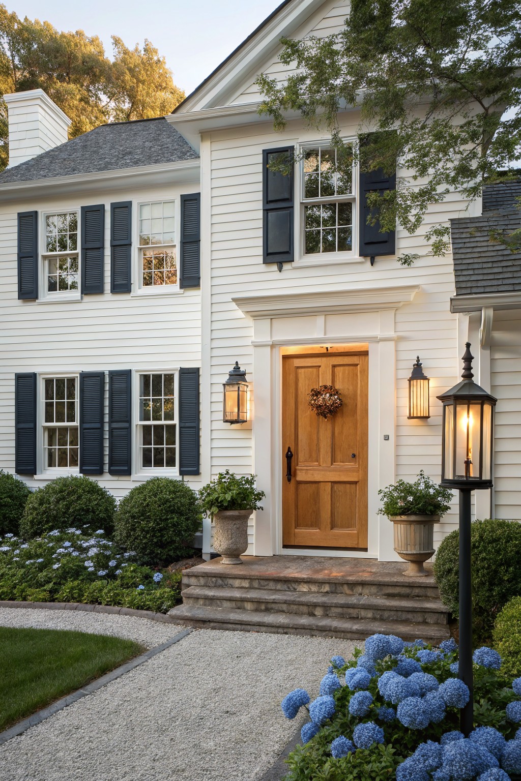 Two-story white clapboard house with black shutters, wooden front door with wreath, lanterns on either side, stone steps, gravel path, boxwood shrubs, and blue hydrangea plantings.