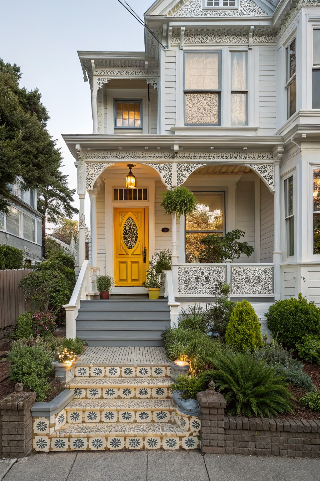 White Victorian house exterior featuring intricate wood trim, a bright yellow front door with decorative glass, a covered porch, gray steps with colorful tile risers, and landscaped plants along the front.