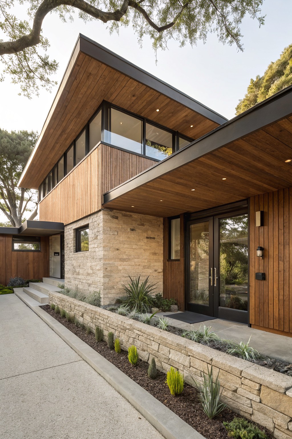 Modern two-story house exterior with vertical brown wood siding on upper levels, beige stone base, black metal roof overhang, large glass entry doors, narrow windows, driveway, and low stone wall with drought-tolerant plants.