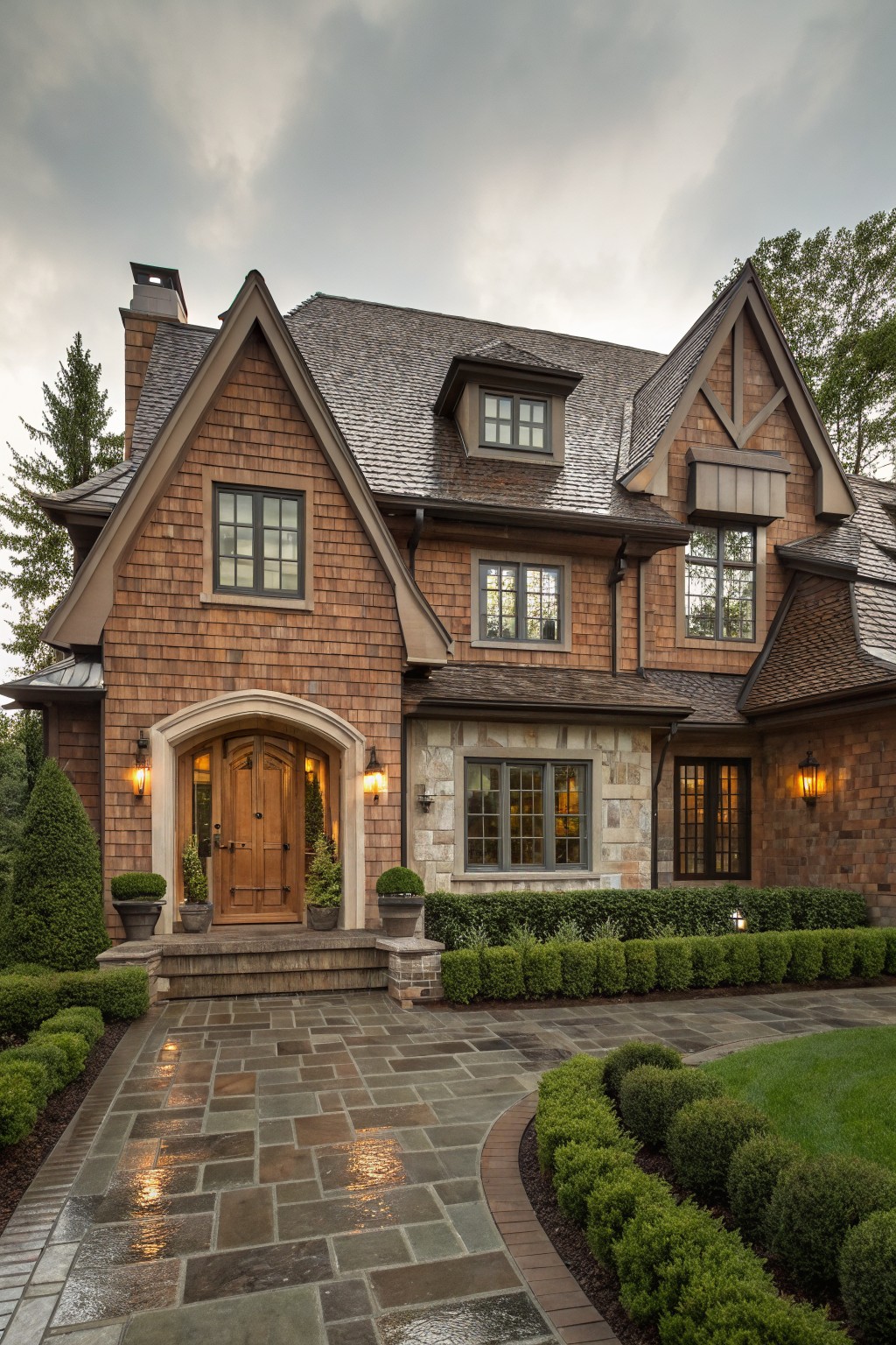 Front view of a two-story house featuring brown shingle siding, multiple gabled roofs, arched wooden entry door flanked by lanterns, stone base accents, and low hedges bordering a slate driveway under overcast skies.