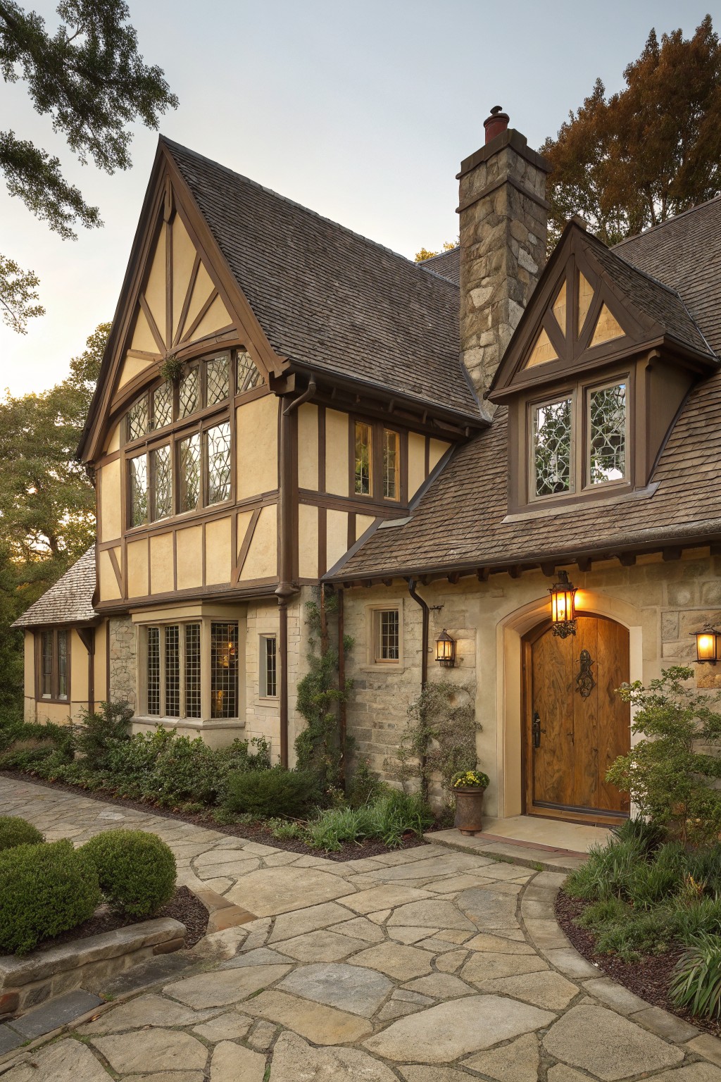 Three-quarter view of a beige stucco Tudor-style house with dark timber framing, stone chimney and entry archway, wooden door, lantern lights, stone pathway, and surrounding shrubs and trees at dusk.