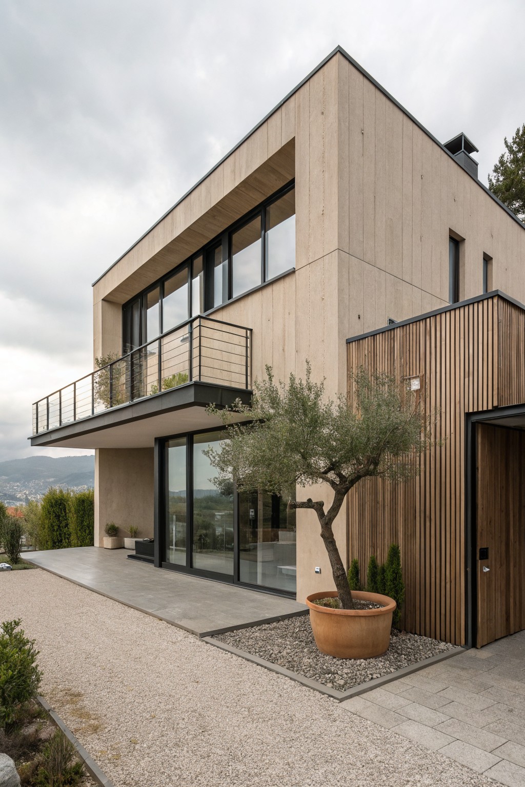 Modern three-story house exterior featuring light brown vertical wood cladding, large black-framed glass windows and doors, a metal balcony railing, and an olive tree in a large terracotta pot beside the entrance on a gravel path.