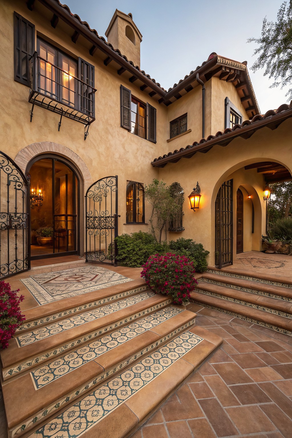 Beige stucco house exterior with terracotta roof tiles, arched entry framed by wrought iron gates, tiled steps leading to a courtyard, and low shrubs and potted flowers nearby.