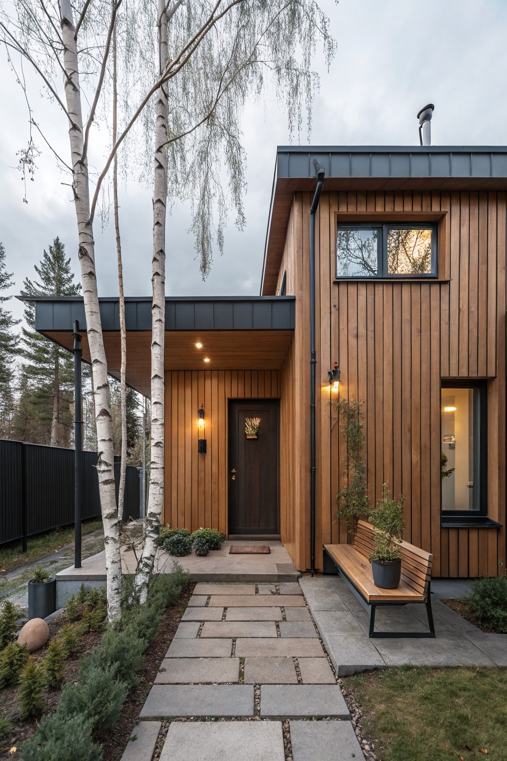 Modern house exterior featuring warm brown vertical wood cladding, black metal roof and accents, wooden front door, built-in bench with plant, stone pathway, and surrounding landscaping with birch trees.