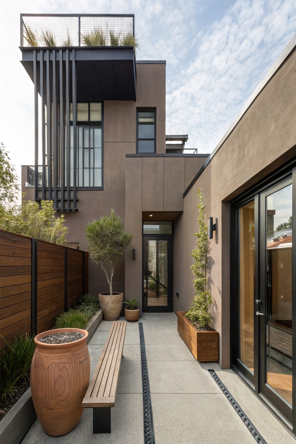 Modern house exterior with taupe stucco walls, black metal vertical accents and window frames, glass entry door flanked by olive trees and large terracotta pots, wooden bench on concrete pathway, wooden fence, and landscaped courtyard.