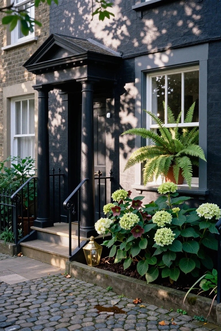 Black-painted brick bungalow exterior with a neoclassical pediment and fluted columns at the front door, white-framed windows, a potted fern in one window, white hydrangeas and dark-leaved plants in the front bed, black railing, brass lantern, and cobblestone path.